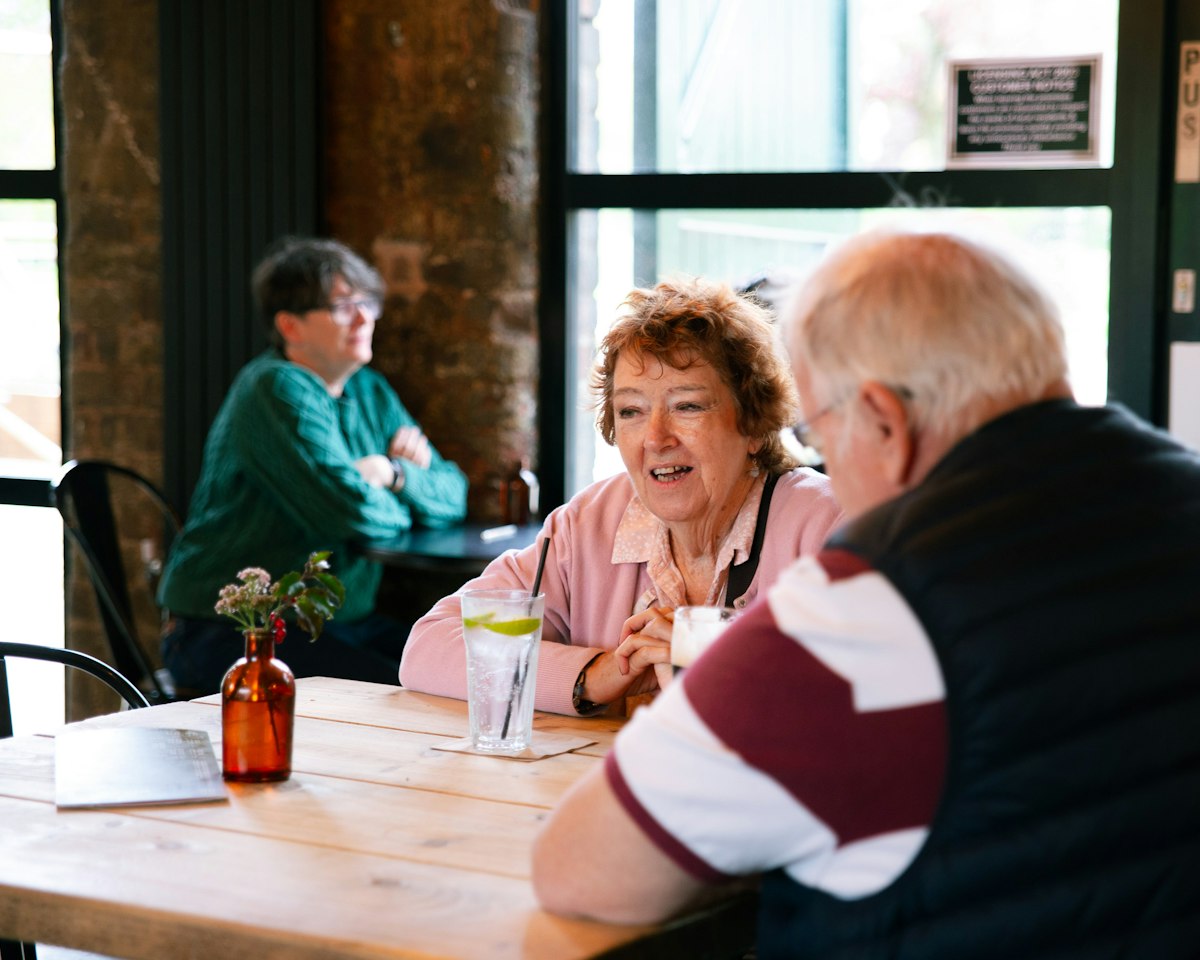 Two couples laugh over drinks at a wooden table in a sunny neighborhood cafe