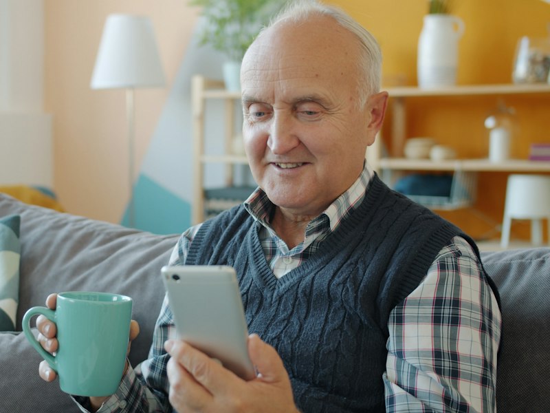 A senior man with a mug of coffee smiles at his phone on the couch