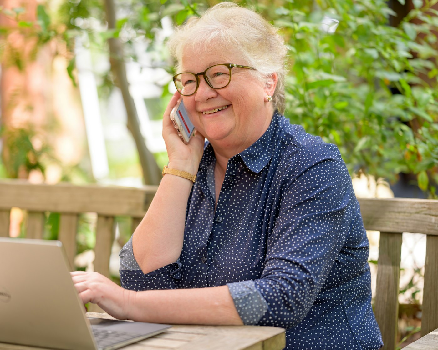 A smiling woman with silver hair talks on her phone at a garden table with her laptop open
