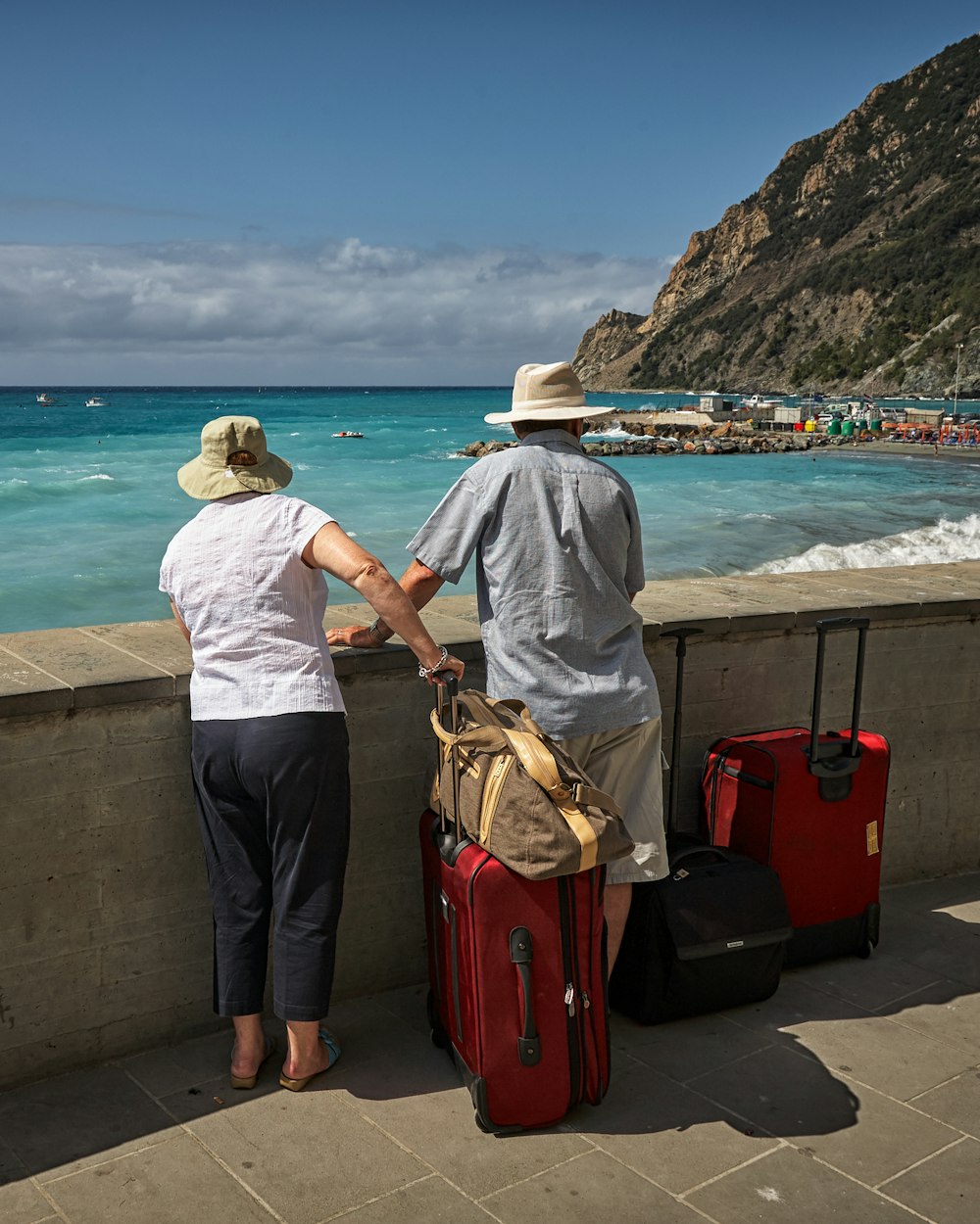 An older couple with suitcases looks out over a turquoise bay from a seaside promenade